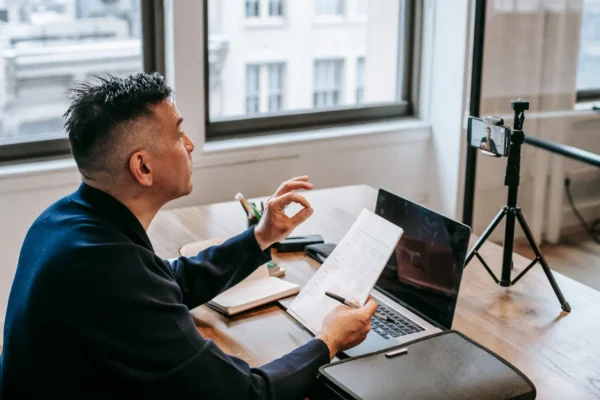 Profissional em escritório usando laptop e papeis para planejar e analisar resultados, enquanto grava conteúdo em câmera com tripé e celular na mesa, ilustrando como melhorar a presença digital de uma empresa em mercados competitivos.