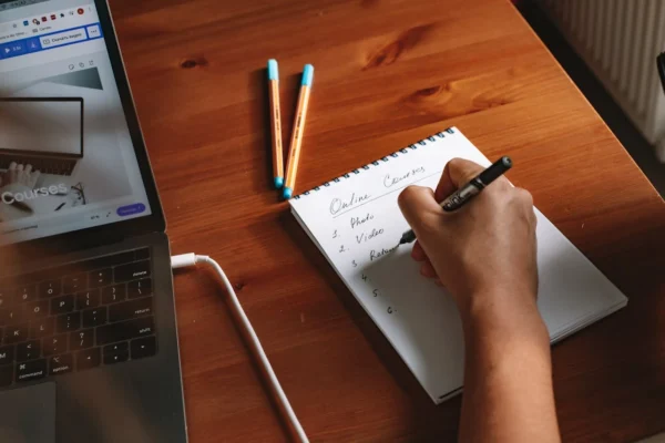 Pessoa fazendo anotações em um caderno sobre cursos online, enquanto um notebook exibe uma plataforma de aprendizagem em um ambiente de trabalho, simbolizando como os cursos online se tornaram essenciais para profissionais autônomos.