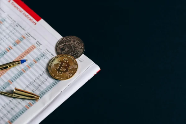 Cenário com moedas digitais e moedas de Bitcoin sobre uma planilha de impostos com caneta ao lado, simbolizando Treine equipe para a nova realidade fiscal e a gestão de dados tributários