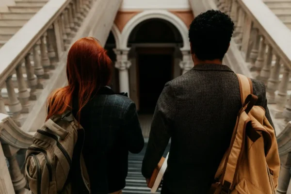 Casal de intercâmbio com mochilas sobe escadas de um prédio antigo, visto por trás, em ambiente urbano, sugerindo planejamento e preparo para estudar fora.