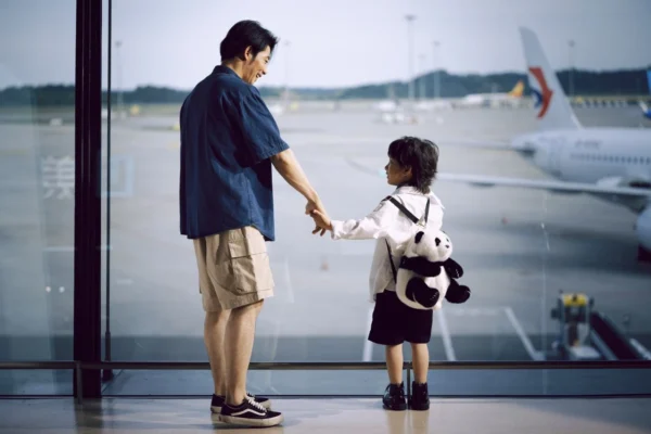 Pai e criança de mãos dadas em área de aeroporto, com um urso de pelúcia, enquanto um avião está ao fundo; imagem sobre Dicas Para Viajar com Crianças: Guia Completo para uma Viagem Tranquila e Divertida.