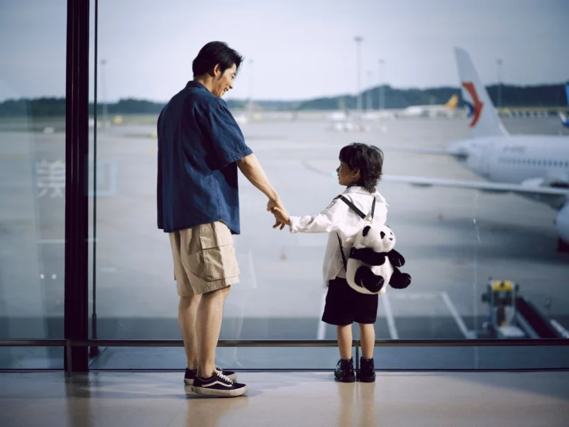 Pai e criança de mãos dadas em área de aeroporto, com um urso de pelúcia, enquanto um avião está ao fundo; imagem sobre Dicas Para Viajar com Crianças: Guia Completo para uma Viagem Tranquila e Divertida.